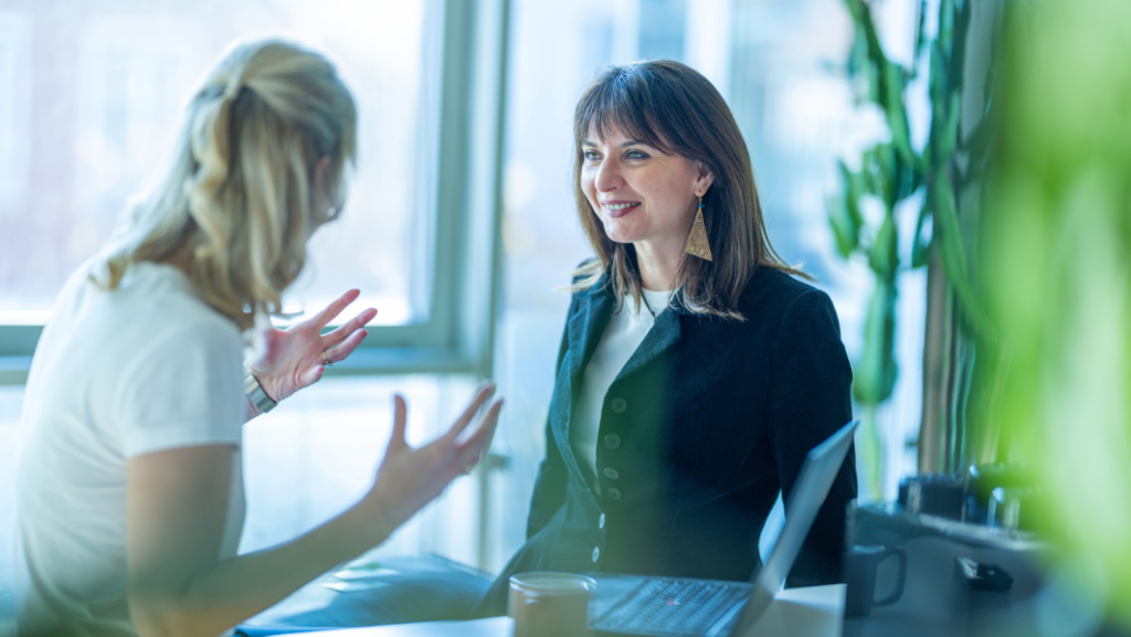 2 Frauen im Gespräch, eine lächelt freundlich, die andere sieht man von hinten