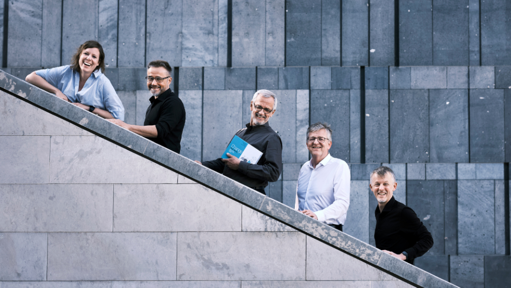 Editorenteam, 5 Personen, auf Stiege, die ins die Kamera lächeln